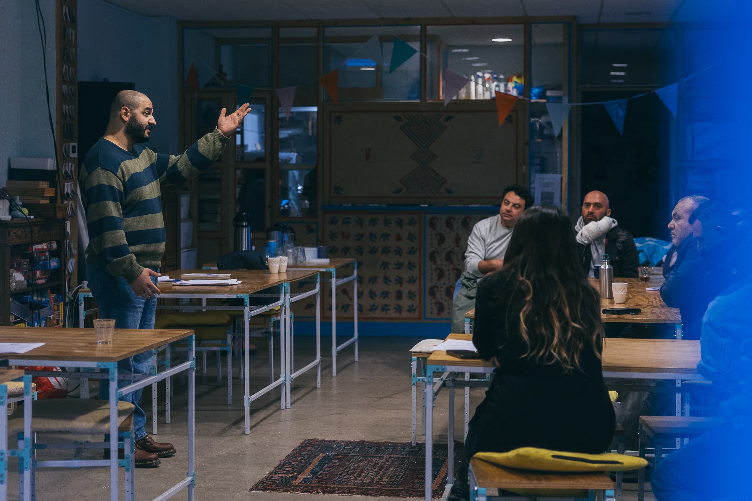 A photo of the storytelling course giver, at De Voorkamer, speaking in from of people (the participants of the workshop) as they are sitting on the tables.