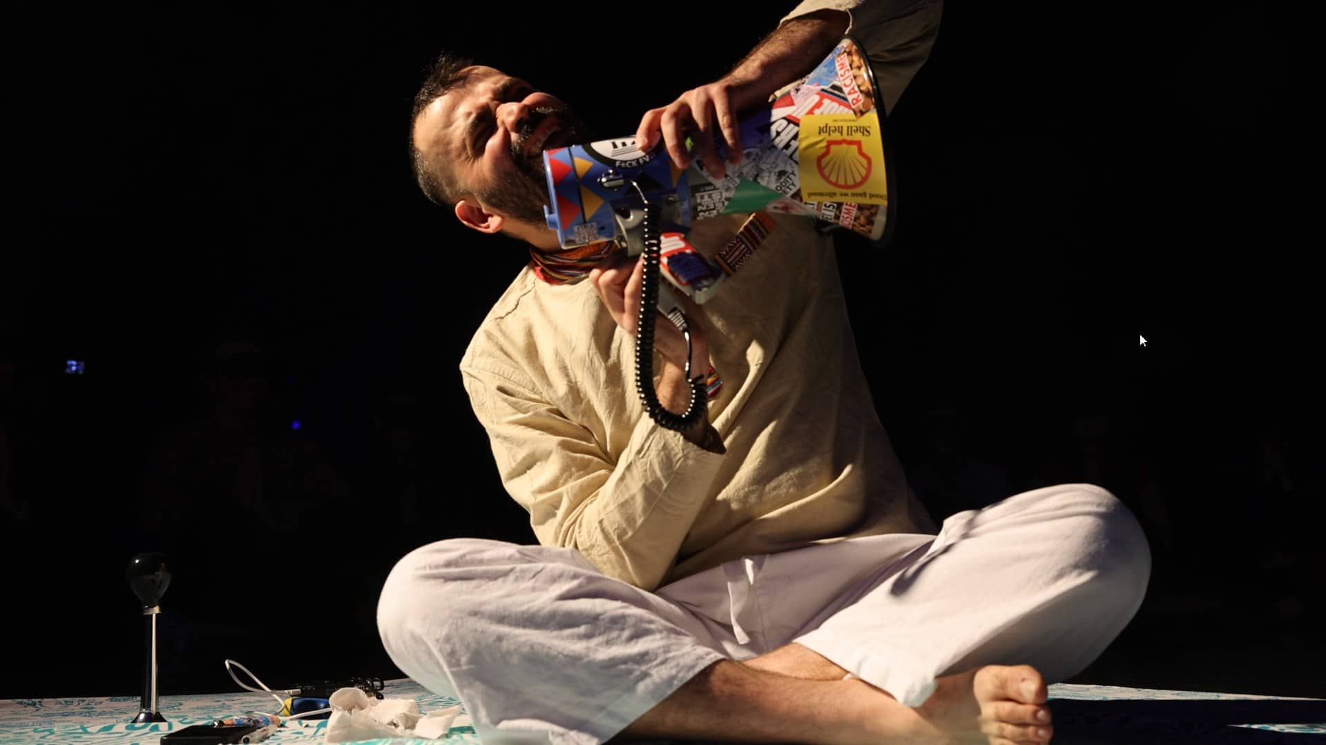 Image of Omid sitting down, crossed legged, on a theatre stage, screaming into a megaphone with stickers on it